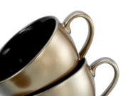 Detailaufnahme aus schräger Seitenperspektive: zwei stapelbare Keramik-Kaffeetassen in Metallic-Gold mit schwarzen Innenflächen und geschwungenen Henkeln vor weißem Hintergrund