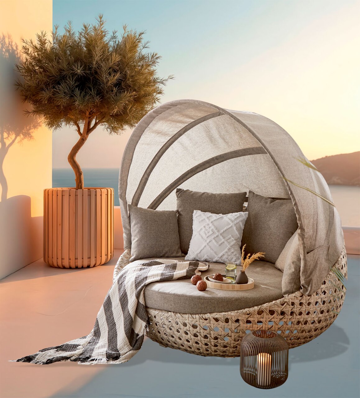 Amatio Tuinschelp DONNA Een beige buitenligbed in ronde rotanlook met een halfronde zonnekap en grijze kussens op een terras aan zee bij zonsondergang, in perspectief vanuit een licht schuin standpunt van rechtsvoor