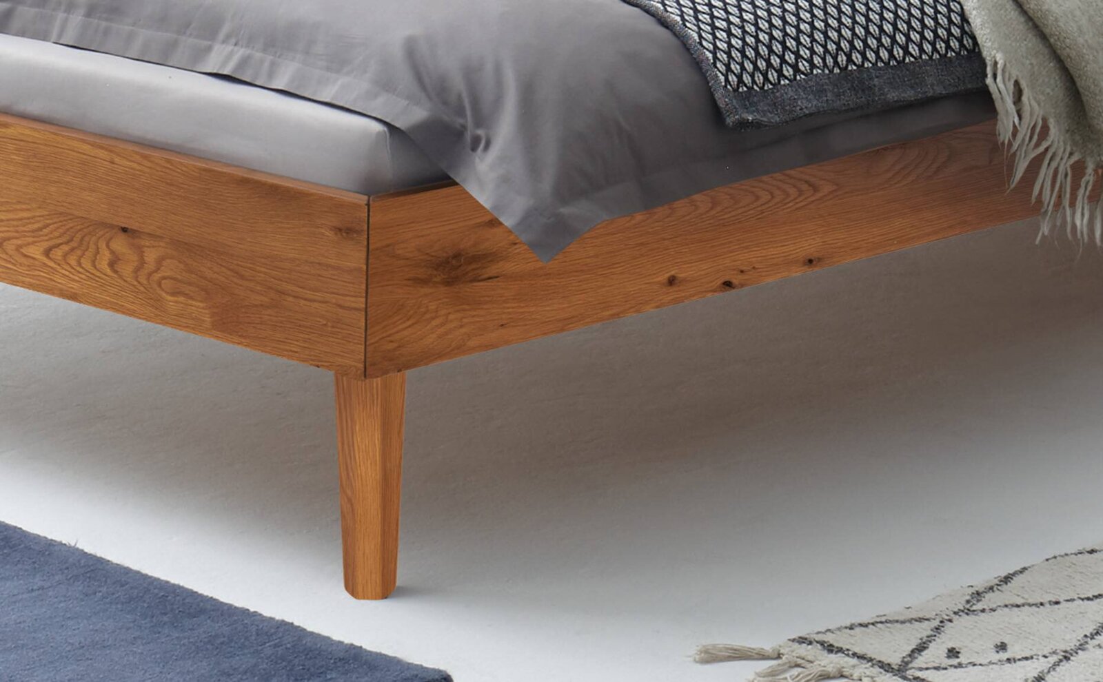 Detailansicht eines modernen Bettrahmens aus massivem Holz mit sichtbarer Maserung und konischem Holzbein, mit grauer Bettwäsche und Decke, aufgenommen aus seitlicher Perspektive von schräg unten.