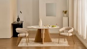 Frontalansicht eines modernen Essbereichs mit rundem Esstisch mit weißer Tischplatte und Holzfuß sowie zwei beigefarbenen Polsterstühlen auf hellem Teppich, dekoriert mit Vase, Gläsern und Obst in einem hellen Raum.