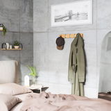 Wandgarderobe aus Holz mit mehreren Haken, an der eine grüne Jacke und eine braune Kappe hängen, in einem modernen Schlafzimmer mit Betonwänden. Perspektive von der Seite.