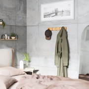 Wandgarderobe aus Holz mit mehreren Haken, an der eine grüne Jacke und eine braune Kappe hängen, in einem modernen Schlafzimmer mit Betonwänden. Perspektive von der Seite.