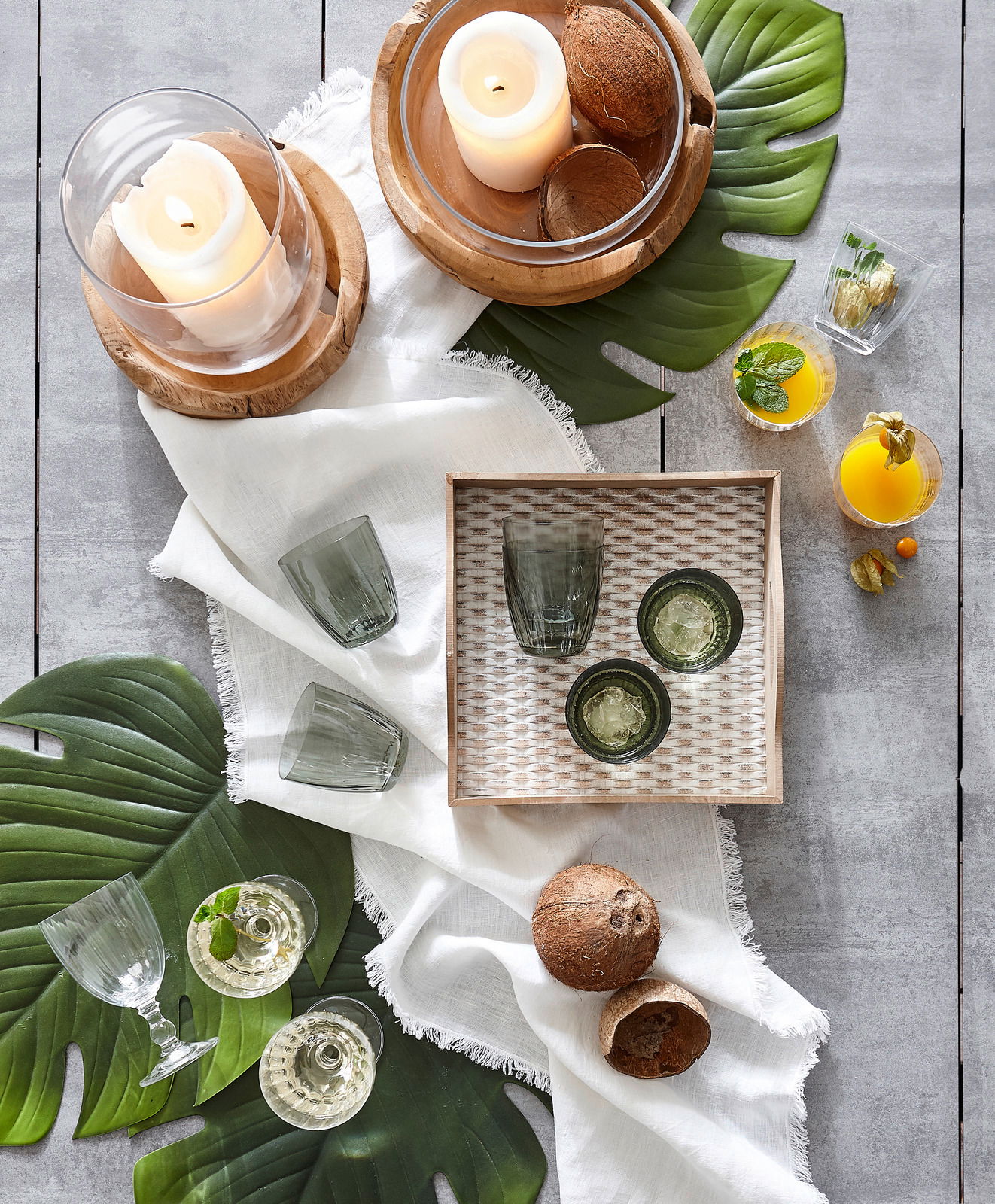 Vogelperspektive auf ein stilvolles Arrangement mit Windlicht aus Teakholz, umgeben von grünen Blättern, Gläsern und exotischen Früchten auf einem hellen Tuch.