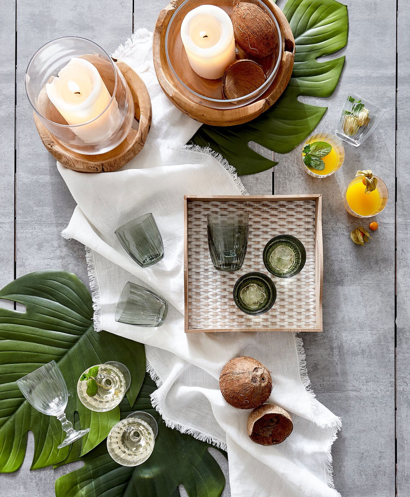 Vogelperspektive auf ein stilvolles Arrangement mit Windlicht aus Teakholz, umgeben von grünen Blättern, Gläsern und exotischen Früchten auf einem hellen Tuch.