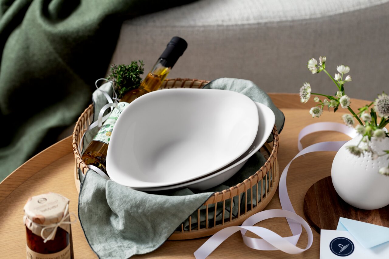 Twee witte pastaschalen in een mand, omringd door een fles olijfolie, kruiden en decoratie, gefotografeerd vanuit vogelperspectief.