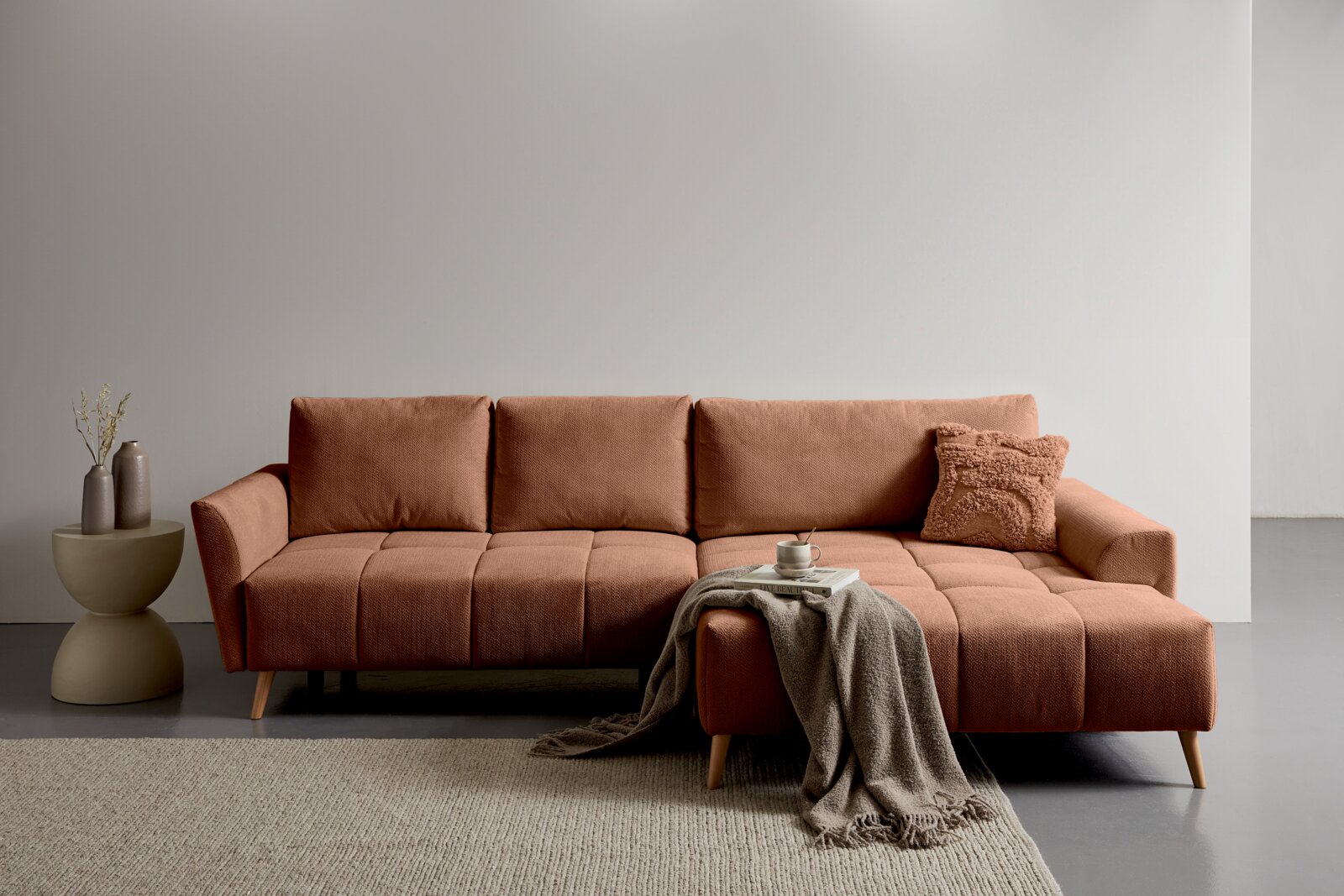 Z2 Ecksofa ZELDA Modernes, terrakottafarbenes Ecksofa mit Chaiselongue rechts, gestepptem Sitz und Holzfüßen; Frontansicht in einem minimalistischen Wohnzimmer mit Beistelltisch, Kissen, Decke und Tasse.