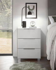 Moderne weiße Nachtkommode mit drei Schubladen, dekoriert mit einer Lampe, einem Wecker und einem Bild, seitliche Perspektive.