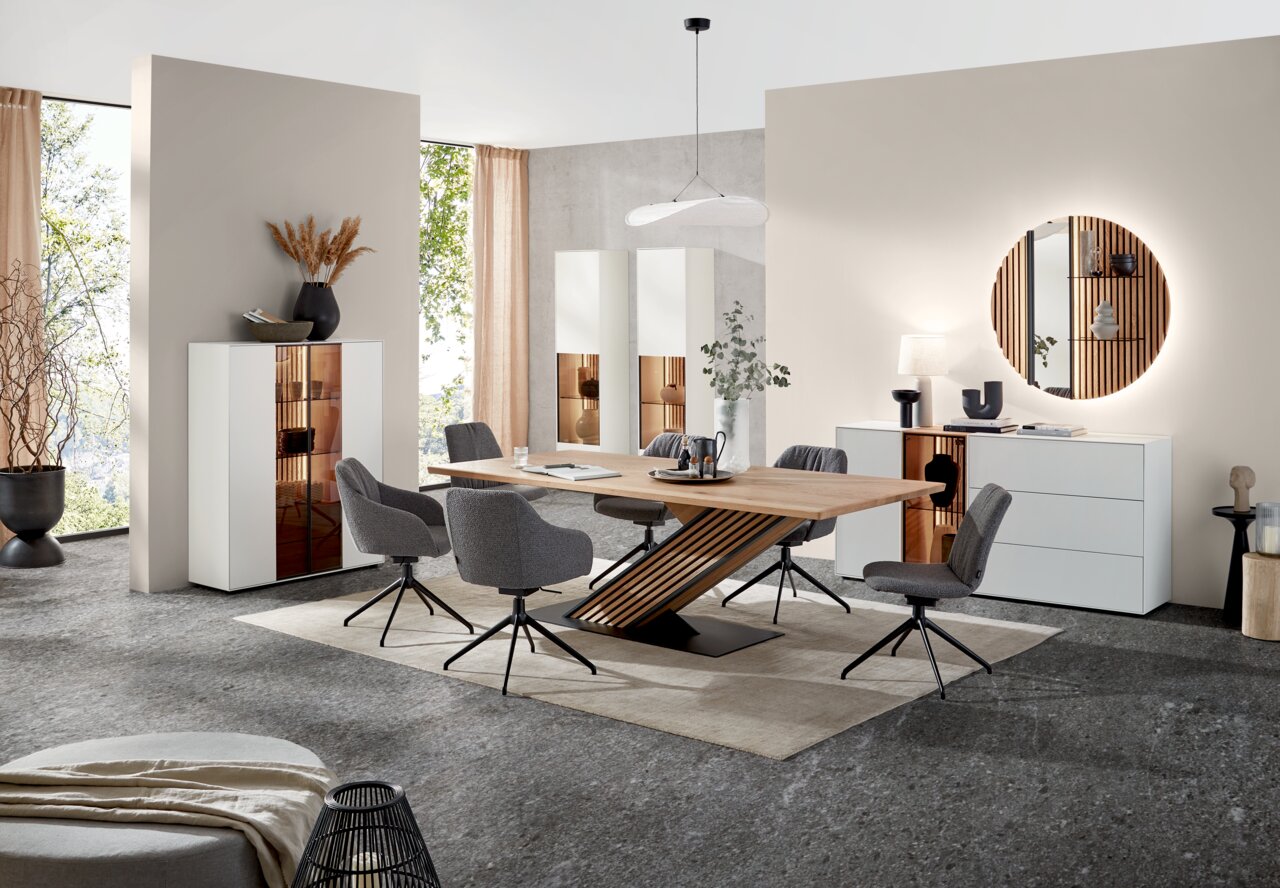 Johann Jakob Esstisch LUCIANO Moderne Esszimmer-Einrichtung mit einem großen Holztisch und grauen Stühlen, flankiert von weißen Vitrinen und einem Sideboard. Perspektive von vorne auf den Tisch und die Stühle.
