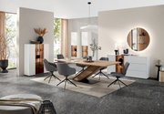 Moderne eetkamerinrichting met een grote houten tafel en grijze stoelen, geflankeerd door witte vitrinekasten en een dressoir. Aanzicht van voren op de tafel en de stoelen.