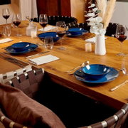 Schräg von oben fotografierter, gedeckter Esstisch aus Holz mit blauem Geschirr, Rotweingläsern, Besteck, Servietten, kleinen Kerzen und weißer Vase mit Eukalyptus und Pampasgras.