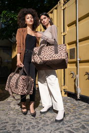 Zwei Frauen stehen vor einem gelben Container und halten Taschen mit Leopardenmuster. Die Perspektive ist frontal.