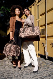 Zwei Frauen stehen vor einem gelben Container und halten Taschen mit Leopardenmuster. Die Perspektive ist frontal.