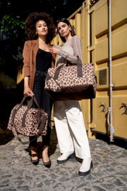 Zwei Frauen stehen vor einem gelben Container und halten Taschen mit Leopardenmuster. Die Perspektive ist frontal.