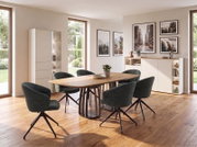 Modernes Esszimmer mit ovalem Holzesstisch und vier dunkelgrünen Drehstühlen auf Holzfußboden, seitliche Perspektive auf das Möbel-Set; im Hintergrund Sideboard, Vitrine und gerahmte Wandbilder bei Tageslicht