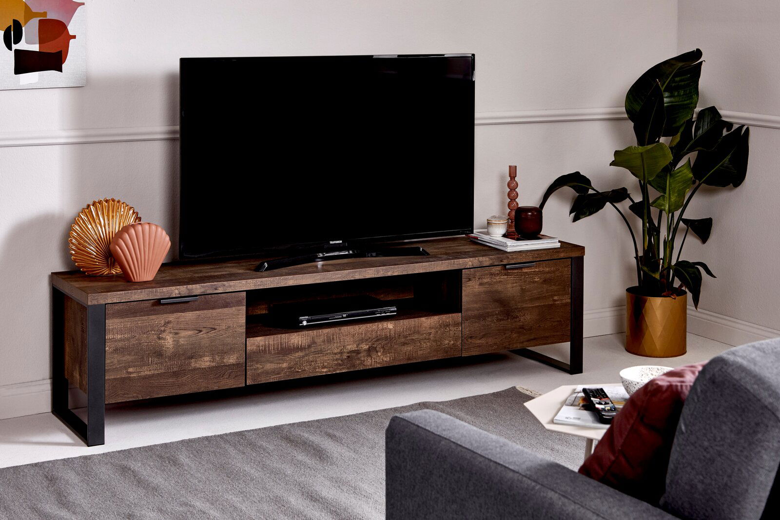 Jahnke TV-Möbel LOOP TV-Möbel aus dunklem Holz mit Metallrahmen, darauf ein großer Fernseher, dekoriert mit Vasen und Pflanzen, Perspektive von vorne.