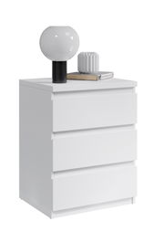 Weiße Nachtkommode mit drei Schubladen, darauf eine moderne Lampe und Dekoration, seitliche Perspektive