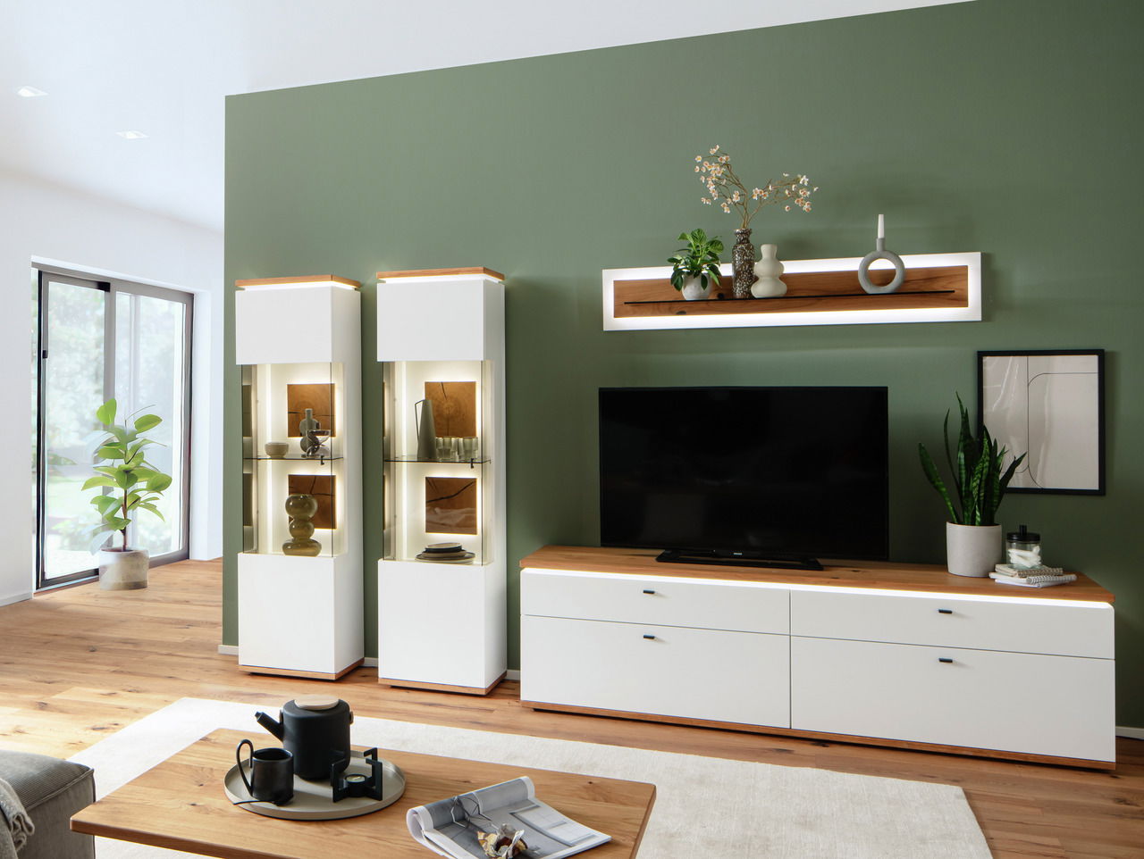 Modernes Wohnzimmer mit grün gestrichener Wand, auf der ein beleuchtetes Wandboard mit Dekorationen angebracht ist. Perspektive: Frontalansicht.