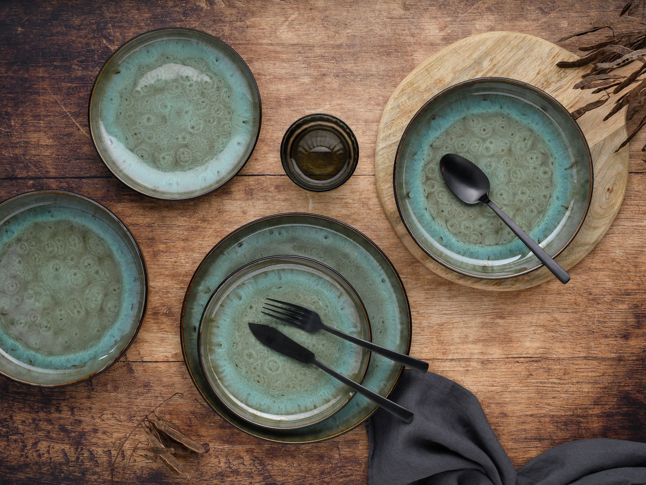 Vogelperspektive auf ein 18-teiliges Tellerset in Grün mit dekorativem Muster, arrangiert auf einem Holztisch mit Besteck und Serviette.