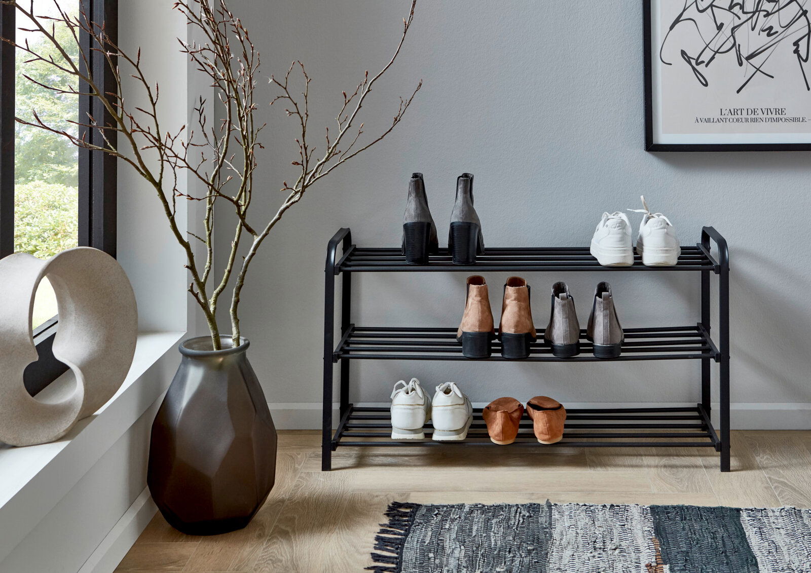 Schwarzes, dreistufiges Schuhregal aus Metall im Flur, frontal aus Augenhöhe fotografiert, mit mehreren Paar Schuhen auf den Ablagen; daneben große Vase mit Zweigen und Fensterlicht.