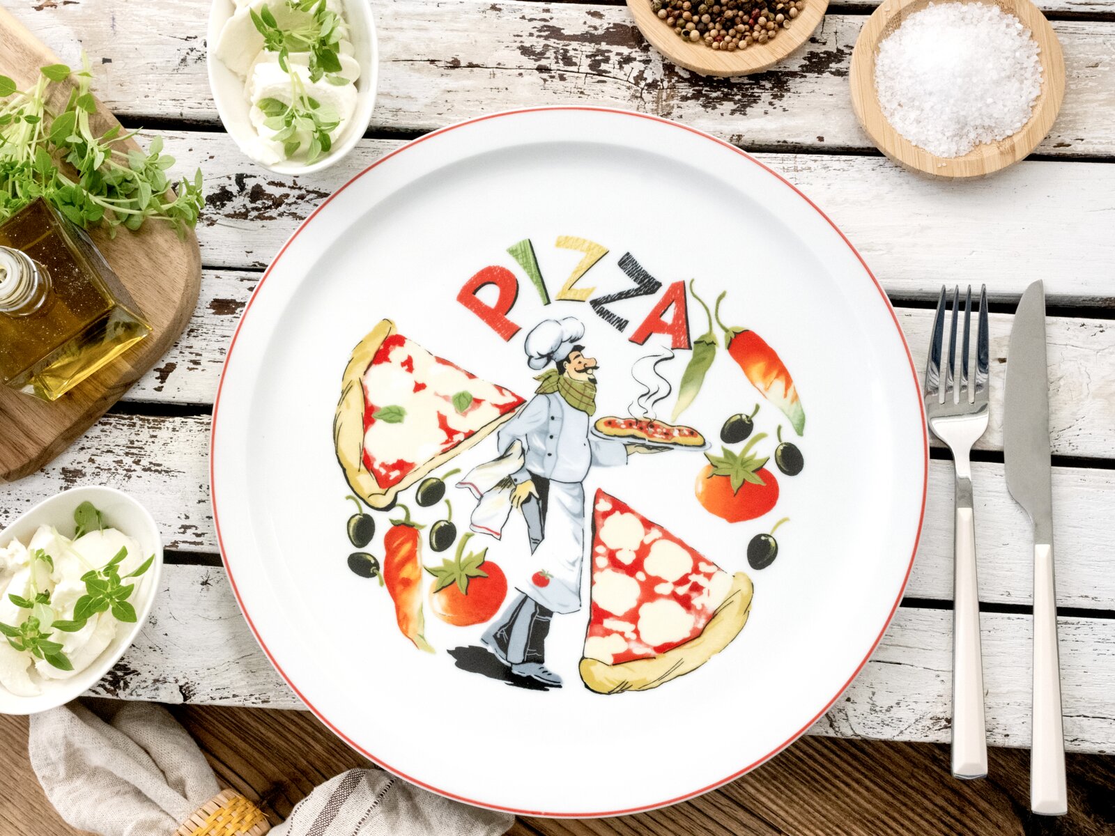 Runder weißer Pizzateller mit rotem Rand und Pizza-Motiv samt Schriftzug „PIZZA“, fotografiert aus der Draufsicht auf rustikalem Holztisch mit Besteck, Olivenöl und Gewürzen.