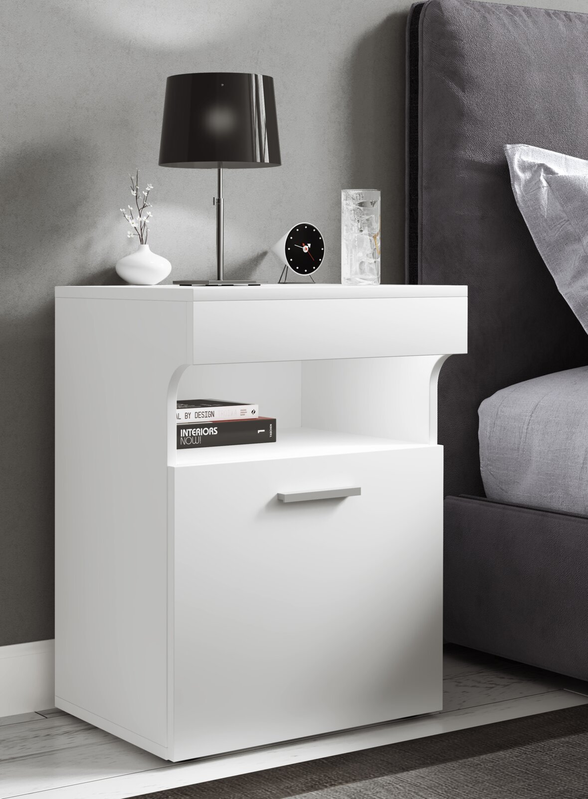 Weißer, moderner Nachttisch mit Schublade und offenem Ablagefach neben einem Bett, darauf Lampe, Uhr, Vase und Glas Ansicht schräg von vorn links.