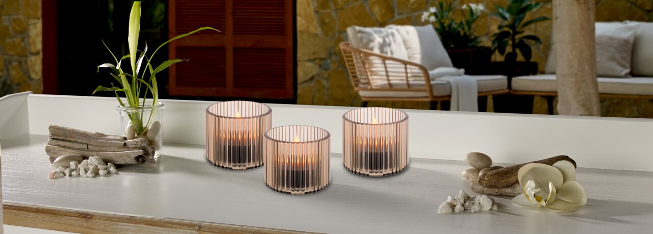 Drei Solarleuchten in eleganten, geriffelten Glasgefäßen auf einem Tisch, umgeben von dekorativen Elementen wie Treibholz und Orchideen, mit einem gemütlichen Außenbereich im Hintergrund. Perspektive: seitlich.