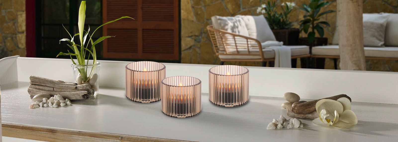 Drei Solarleuchten in eleganten, geriffelten Glasgefäßen auf einem Tisch, umgeben von dekorativen Elementen wie Treibholz und Orchideen, mit einem gemütlichen Außenbereich im Hintergrund. Perspektive: seitlich.