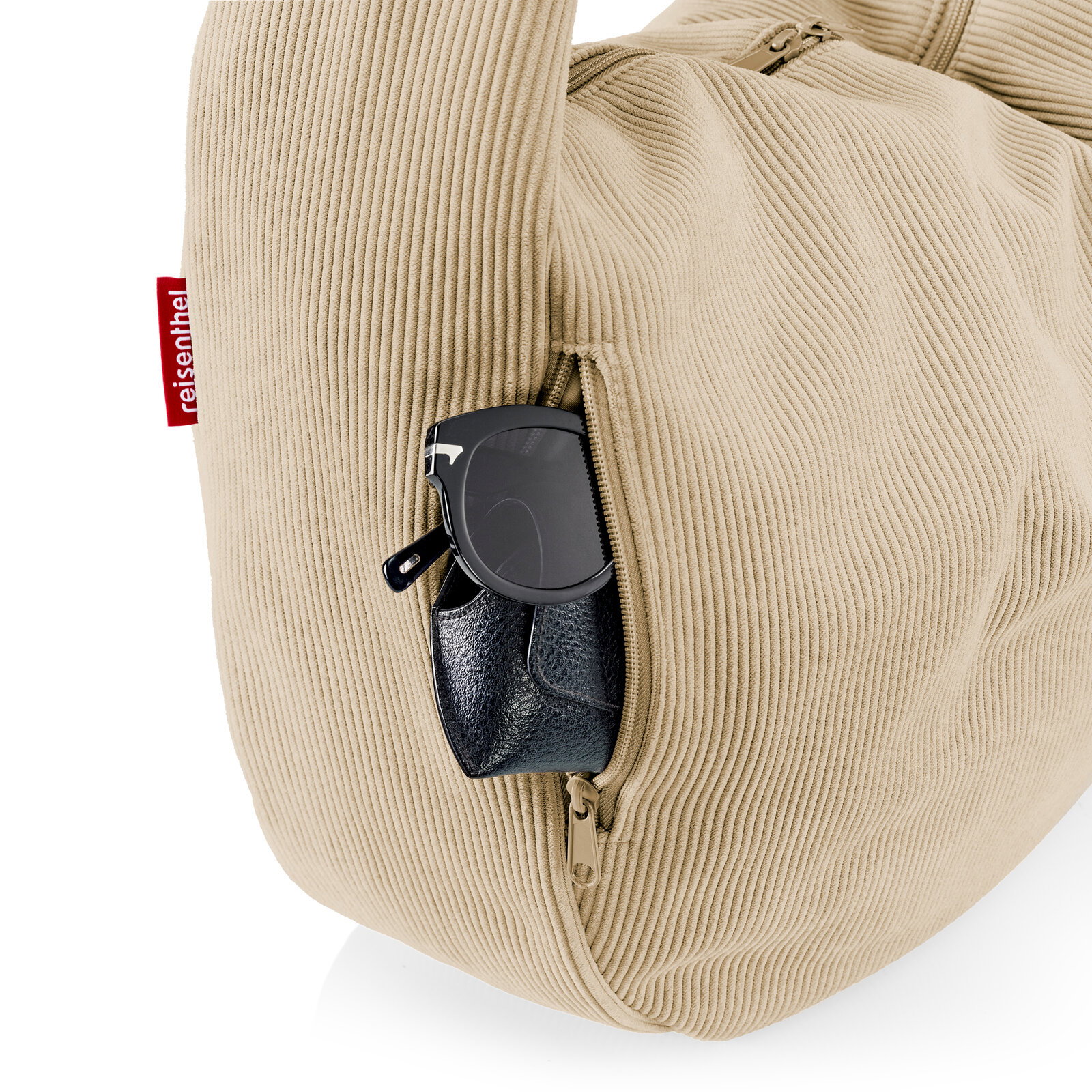 Beige Cord-Reisetasche mit seitlicher Reißverschlusstasche, aus der Sonnenbrille und schwarzes Etui herausragen, Nahaufnahme in seitlicher Perspektive.