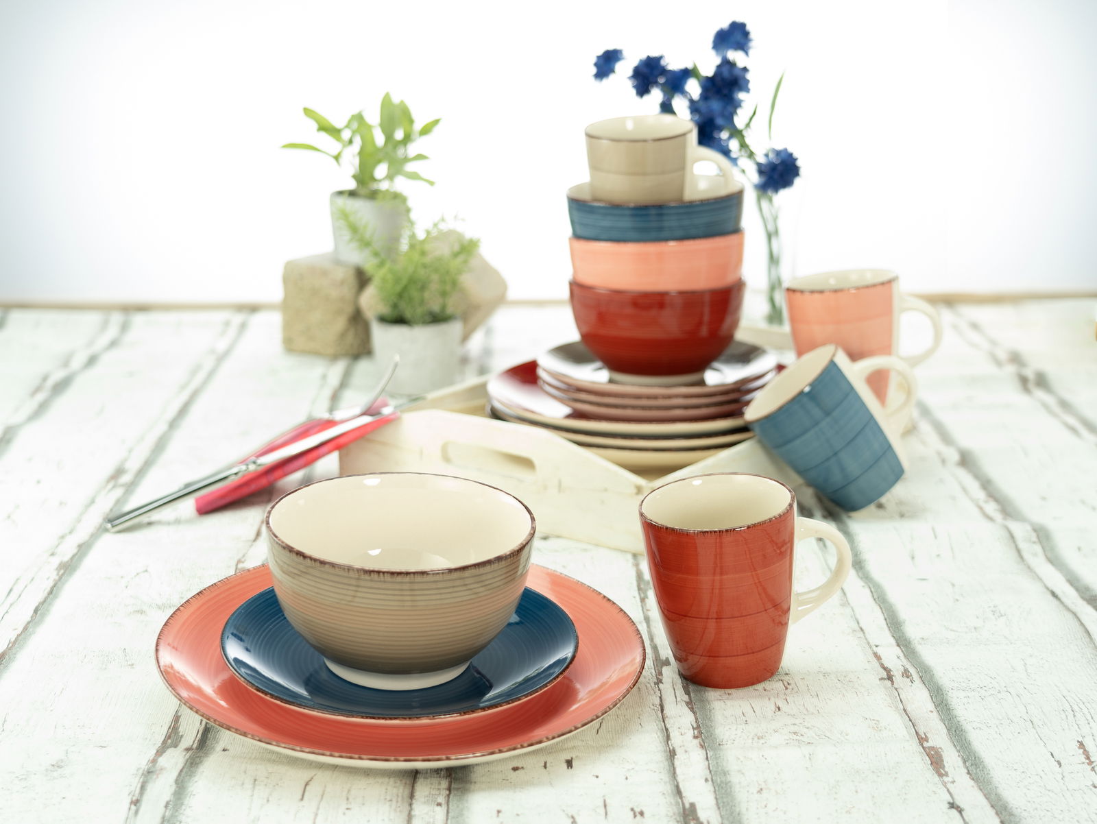 Kombiservice 16-tlg. in verschiedenen Farben, bestehend aus Tellern, Schalen und Tassen, auf einem rustikalen Holztisch arrangiert. Perspektive von vorne.