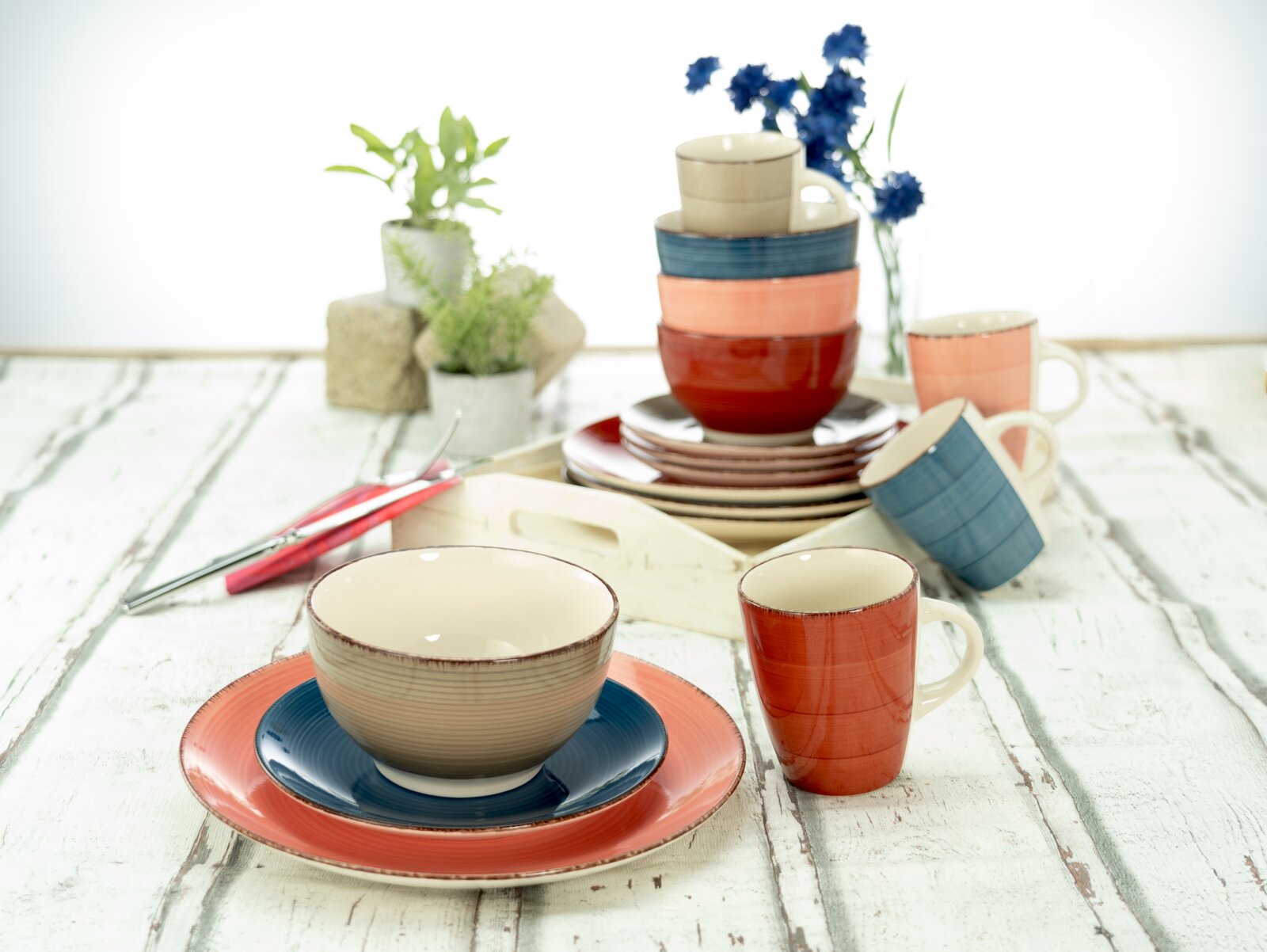 Kombiservice 16-tlg. in verschiedenen Farben, bestehend aus Tellern, Schalen und Tassen, auf einem rustikalen Holztisch arrangiert. Perspektive von vorne.