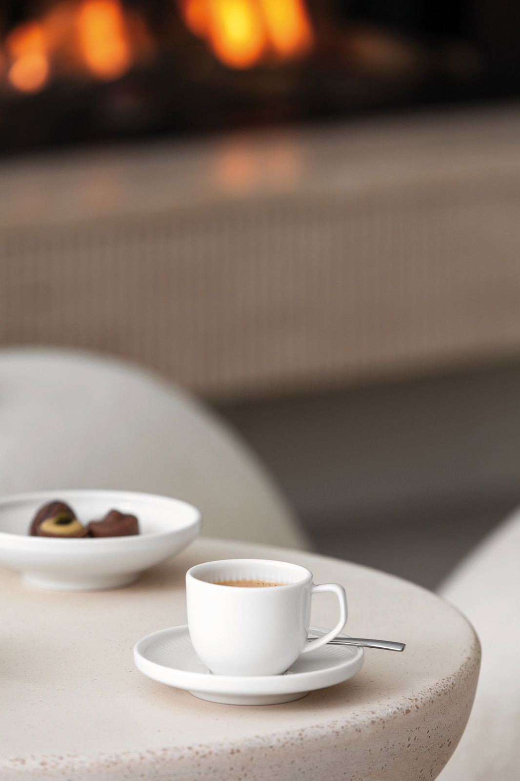 Weiße Espressoobertasse Afina 50ml auf einem Tisch, mit einem Löffel auf der Untertasse, im Hintergrund ein Teller mit Pralinen und ein unscharfer Kamin.