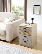 Rollcontainer mit drei Schubladen, aus hellem Holz und Metall, neben einem Sofa in einem Wohnzimmer platziert. Perspektive von vorne rechts.