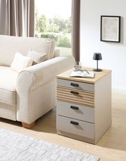 Rollcontainer mit drei Schubladen, aus hellem Holz und Metall, neben einem Sofa in einem Wohnzimmer platziert. Perspektive von vorne rechts.