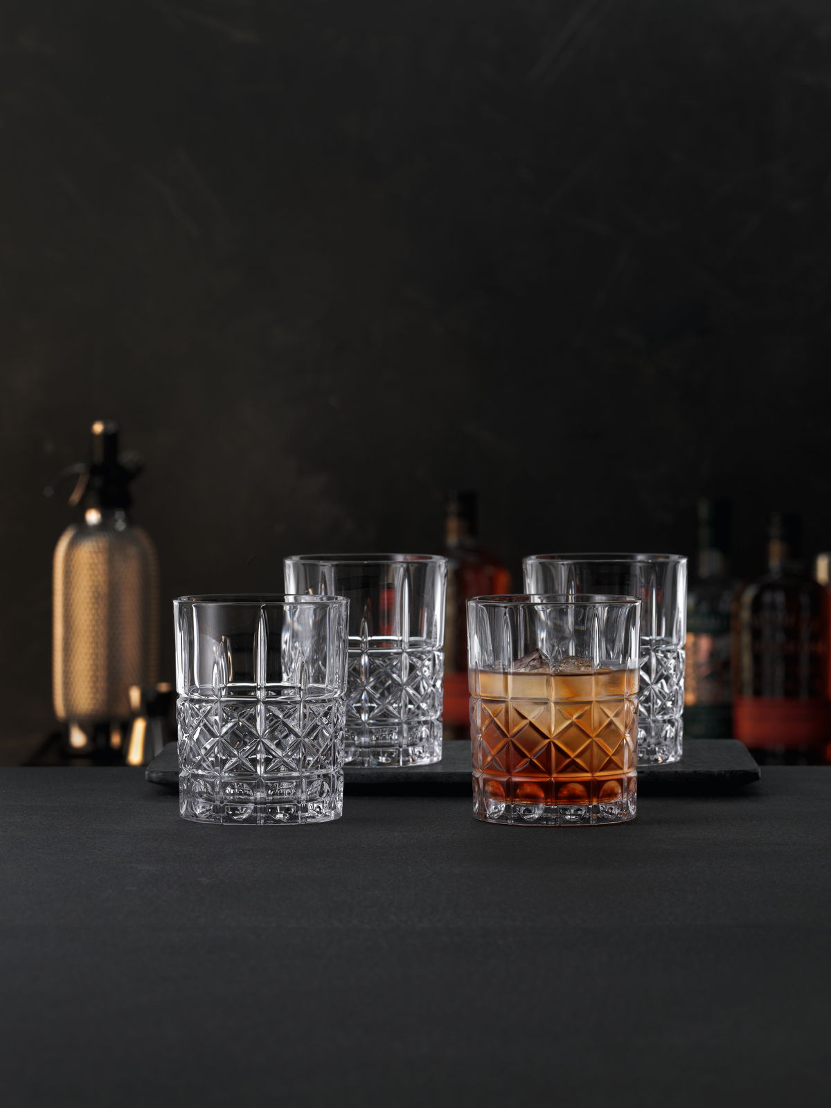 Vier elegante Kristallgläser mit feinem Schliffmuster, eines davon mit Getränk gefüllt, auf einer dunklen Oberfläche in einer stilvollen Bar-Atmosphäre, seitliche Perspektive.