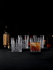 Vier elegante Kristallgläser mit feinem Schliffmuster, eines davon mit Getränk gefüllt, auf einer dunklen Oberfläche in einer stilvollen Bar-Atmosphäre, seitliche Perspektive.