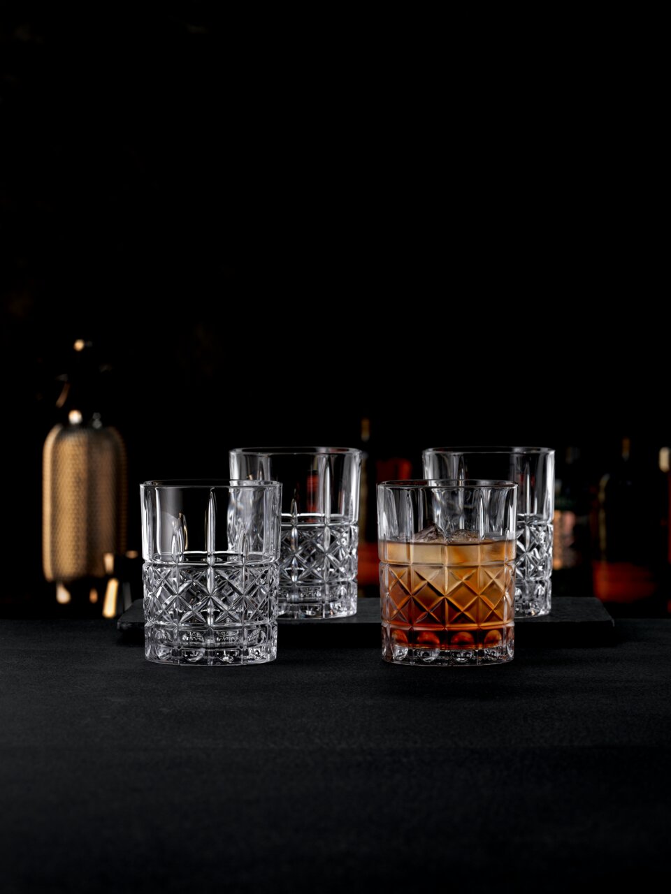 Vier elegante Kristallgläser mit feinem Schliffmuster, eines davon mit Getränk gefüllt, auf einer dunklen Oberfläche in einer stilvollen Bar-Atmosphäre, seitliche Perspektive.