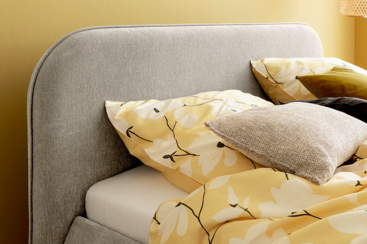 Nahaufnahme eines grauen Polsterbettes mit abgerundetem Kopfteil, dekoriert mit gelber Bettwäsche mit Blumenmuster und mehreren Kissen, seitliche Perspektive.