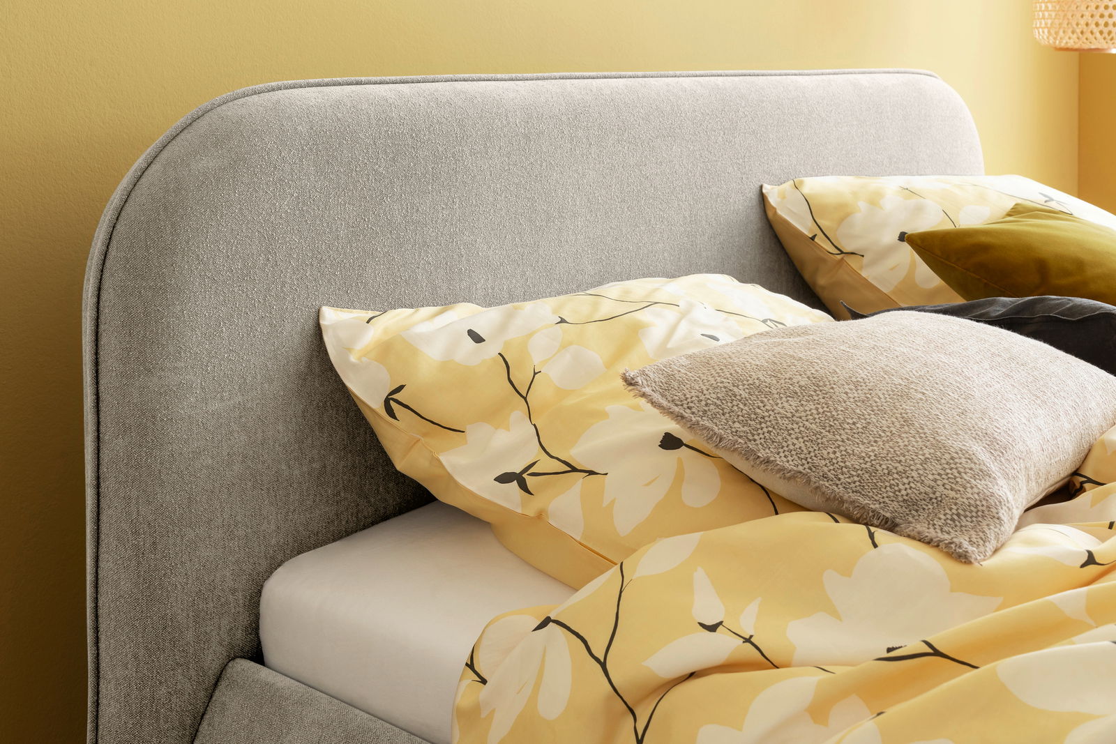 Nahaufnahme eines grauen Polsterbettes mit abgerundetem Kopfteil, dekoriert mit gelber Bettwäsche mit Blumenmuster und mehreren Kissen, seitliche Perspektive.