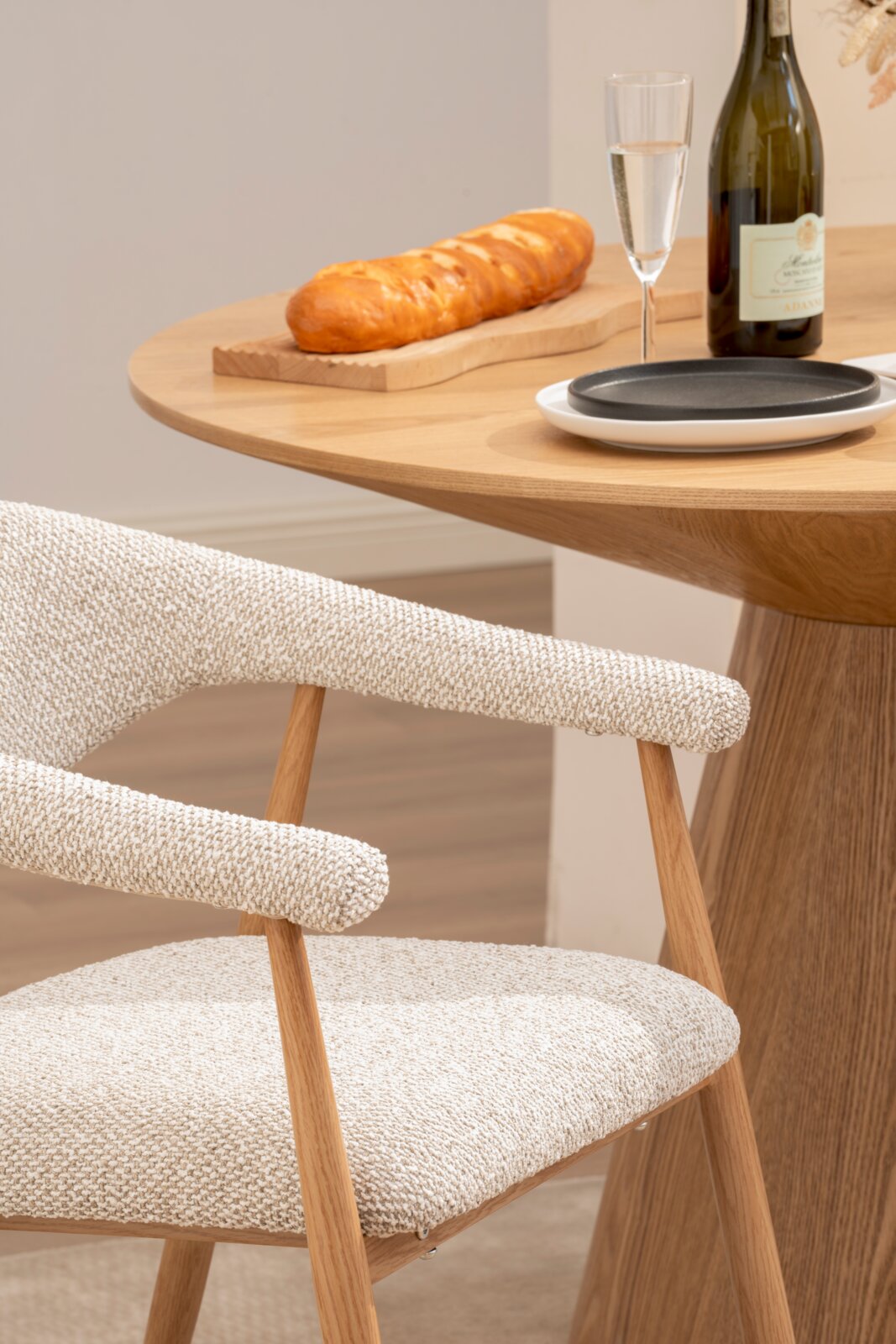 Beiger gepolsterter Esszimmerstuhl mit Holzgestell neben rundem Holztisch mit Brot auf Schneidebrett, Sektglas und Flasche, schräg von der Seite aufgenommen.