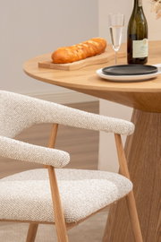 Beiger gepolsterter Esszimmerstuhl mit Holzgestell neben rundem Holztisch mit Brot auf Schneidebrett, Sektglas und Flasche, schräg von der Seite aufgenommen.