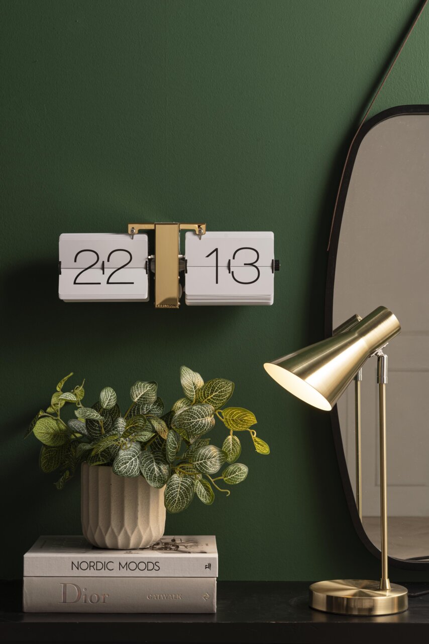 Een flip-klok zonder behuizing in wit en messing, bevestigd aan een groene muur, met op de voorgrond een moderne tafellamp en een plantenarrangement.