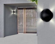 Moderne LED-Außenwandleuchte in schwarzem Design, montiert an einer grauen Wand neben einer Glastür, mit Lichtstrahlen in alle Richtungen.