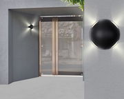 Moderne LED-Außenwandleuchte in schwarzem Design, montiert an einer grauen Wand neben einer Glastür, mit Lichtstrahlen in alle Richtungen.