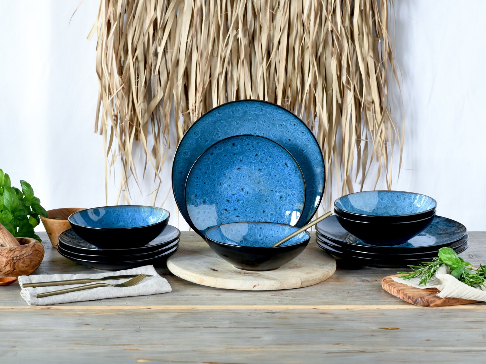 Blau-schwarzes Keramik-Geschirrset mit Tellern und Schalen auf Holztisch dekoriert mit Kräutern, Frontalansicht auf das Arrangement
