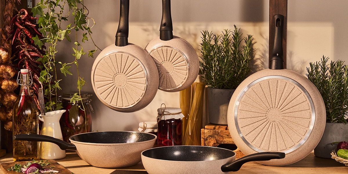 BALLARINI Bratpfanne LEDRO Bratpfannen in einer Küche, aufgehängt und auf einer Arbeitsfläche stehend, mit Pflanzen und Gewürzen im Hintergrund. Perspektive von vorne.