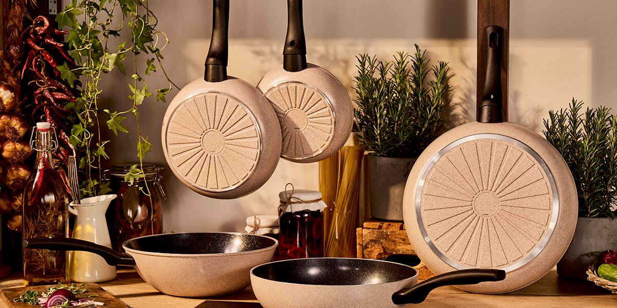 Bratpfannen in einer Küche, aufgehängt und auf einer Arbeitsfläche stehend, mit Pflanzen und Gewürzen im Hintergrund. Perspektive von vorne.