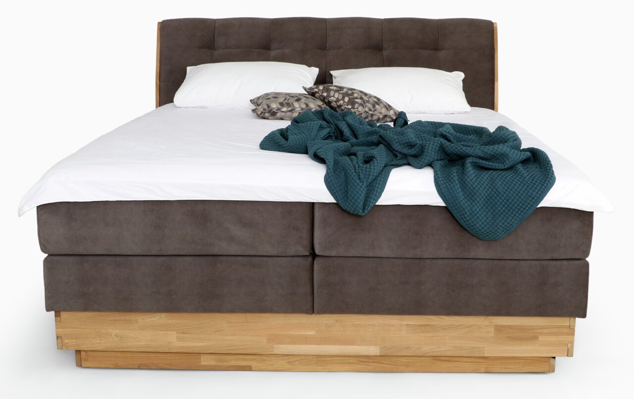 Frontale Ansicht eines Boxspringbetts mit braunem Stoffbezug, zwei weißen Kissen und einer blauen Decke.