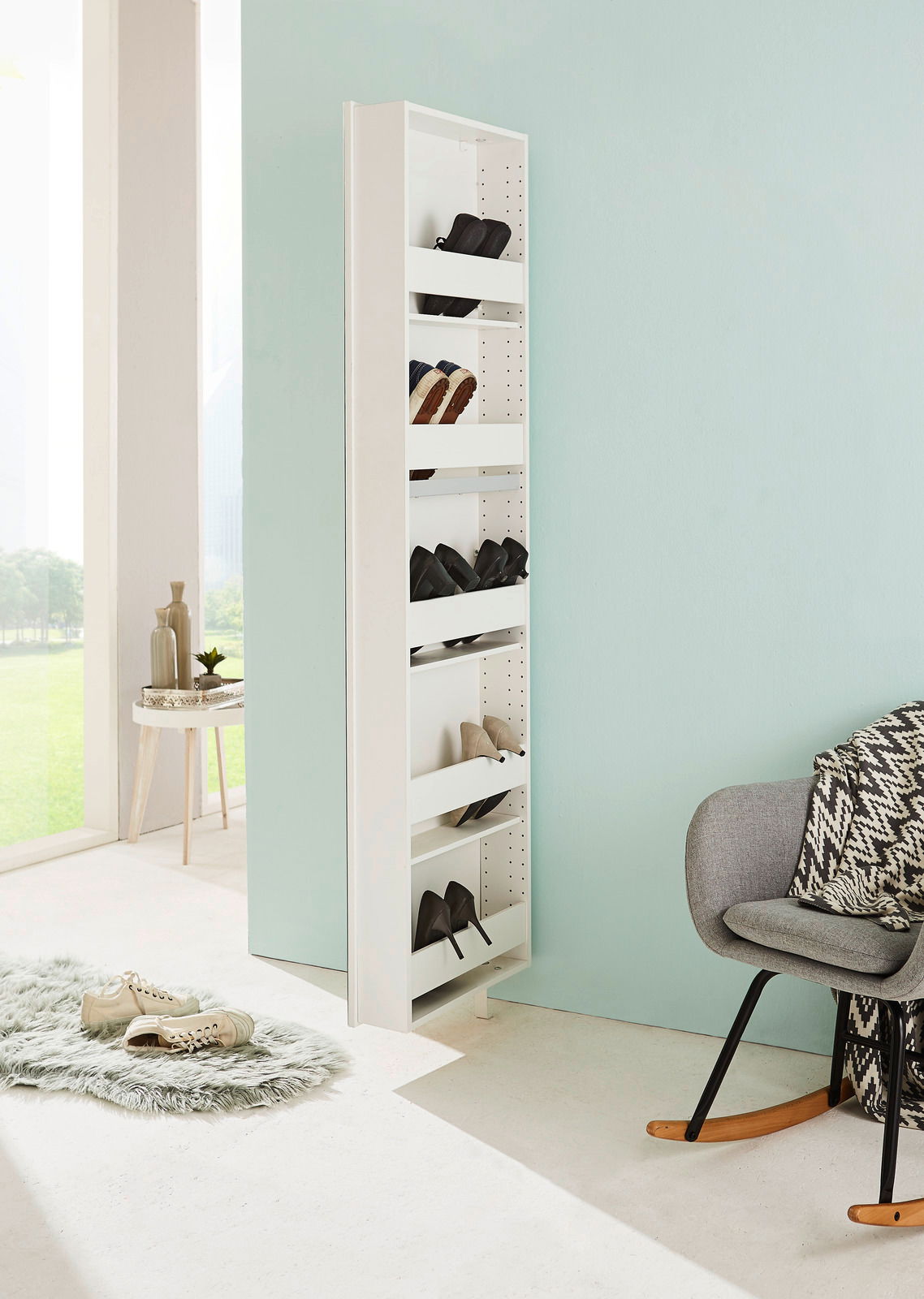 Schmaler, weißer Drehschuhschrank mit mehreren Fächern, gefüllt mit Schuhen, in einem modernen, hellen Raum mit Blick auf eine grüne Landschaft. Perspektive von der Seite.