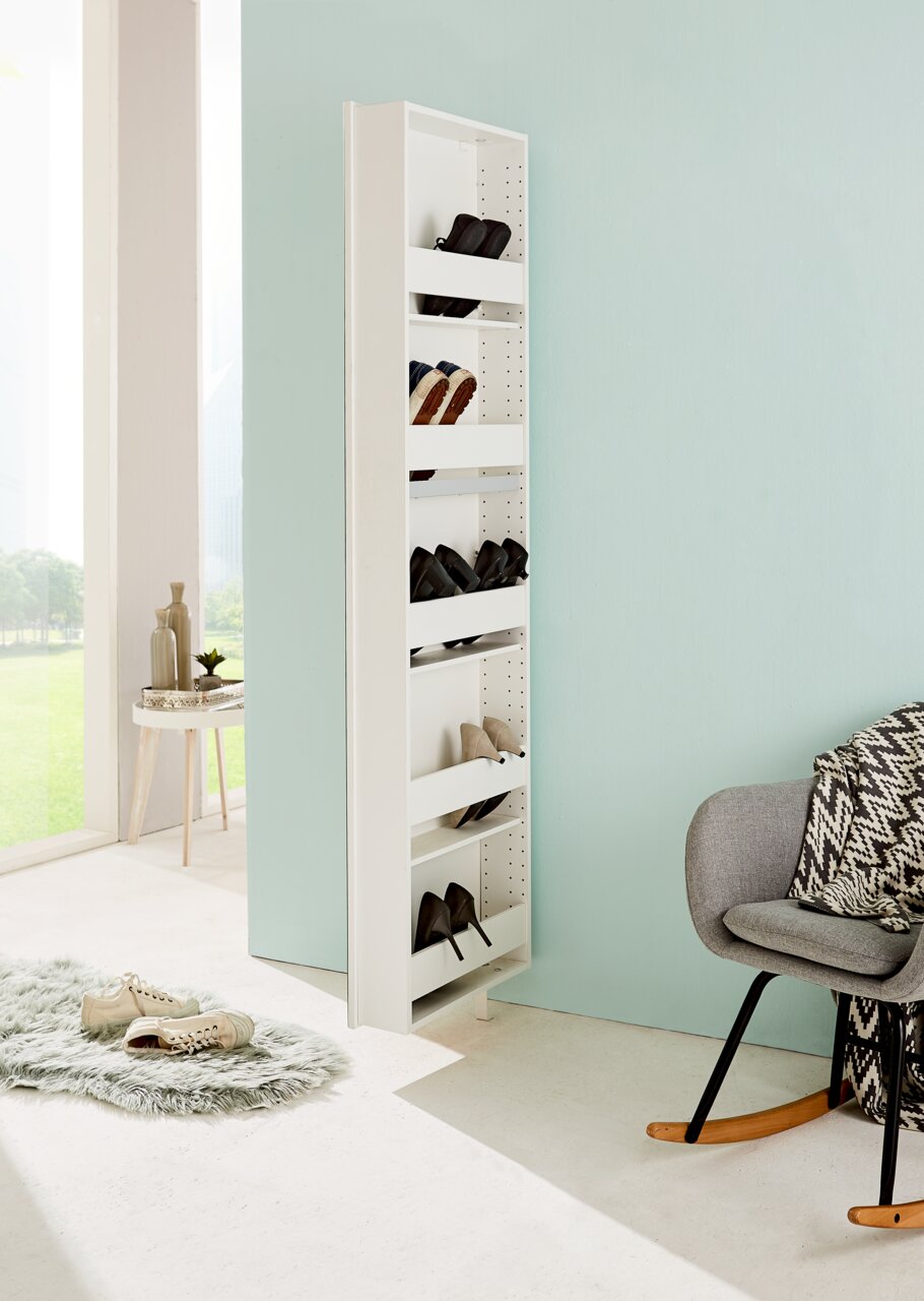 Schmaler, weißer Drehschuhschrank mit mehreren Fächern, gefüllt mit Schuhen, in einem modernen, hellen Raum mit Blick auf eine grüne Landschaft. Perspektive von der Seite.