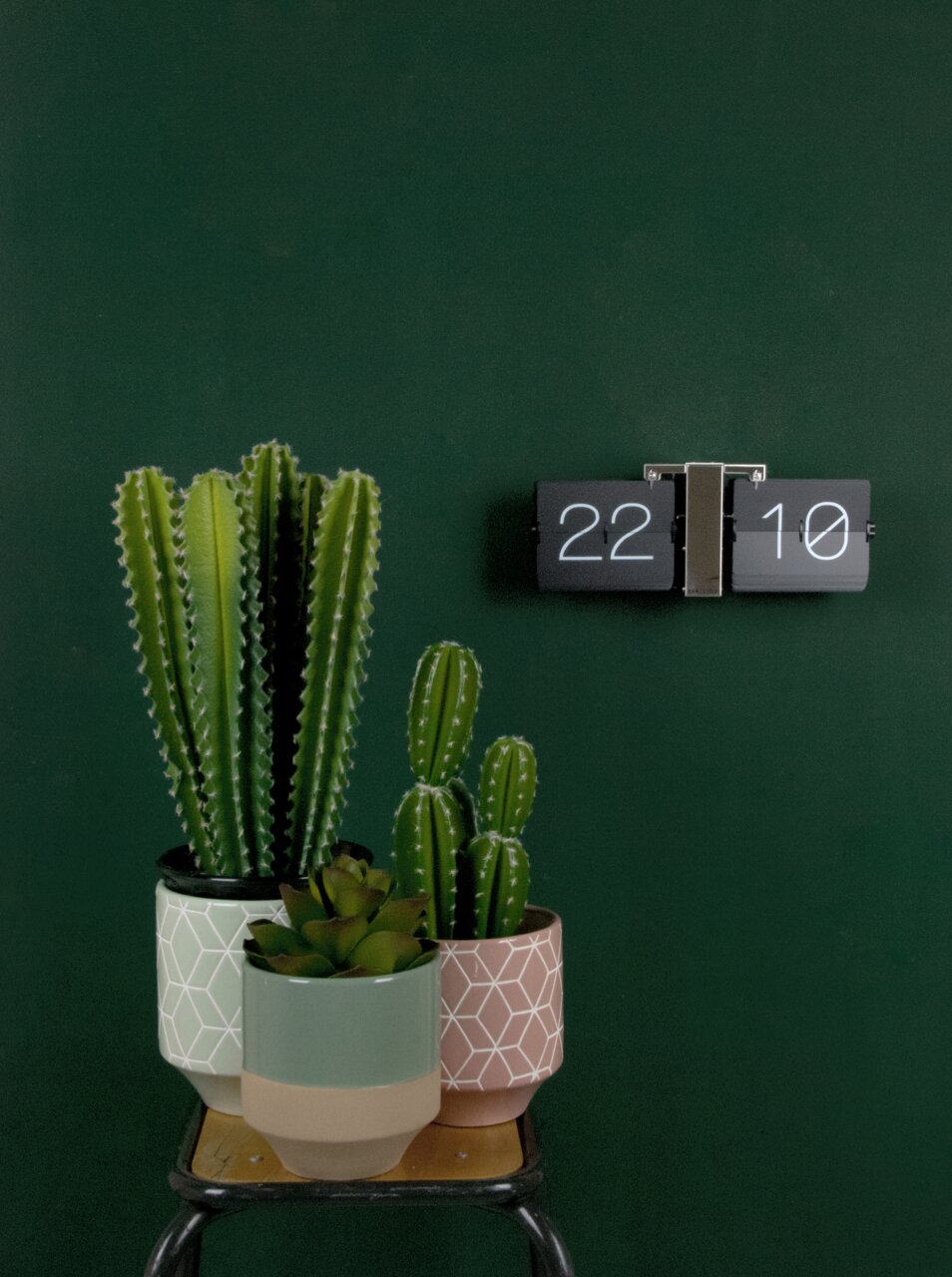 Een moderne, zwarte klok van KARLSSON tegen een groene muur, naast decoratieve cactussen in stijlvolle potten.